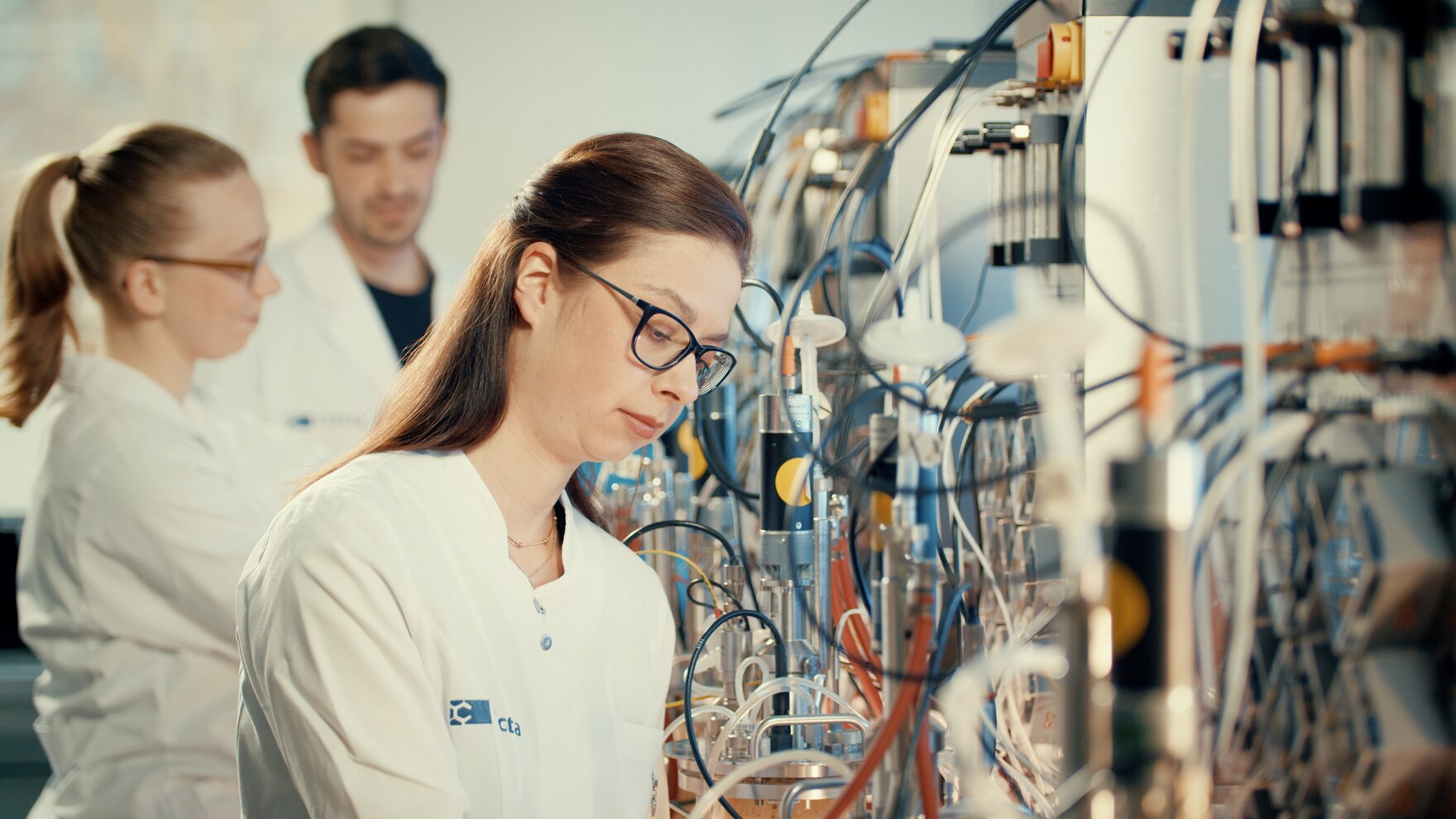 Drei Personen in weißen Kitteln stehen in einem Labor an Geräten.