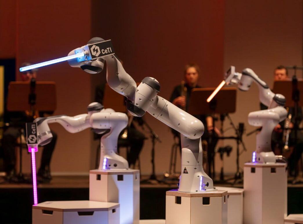 Roboterarme mit leuchtenden Stäben, im Hintergrund sitzen Musiker