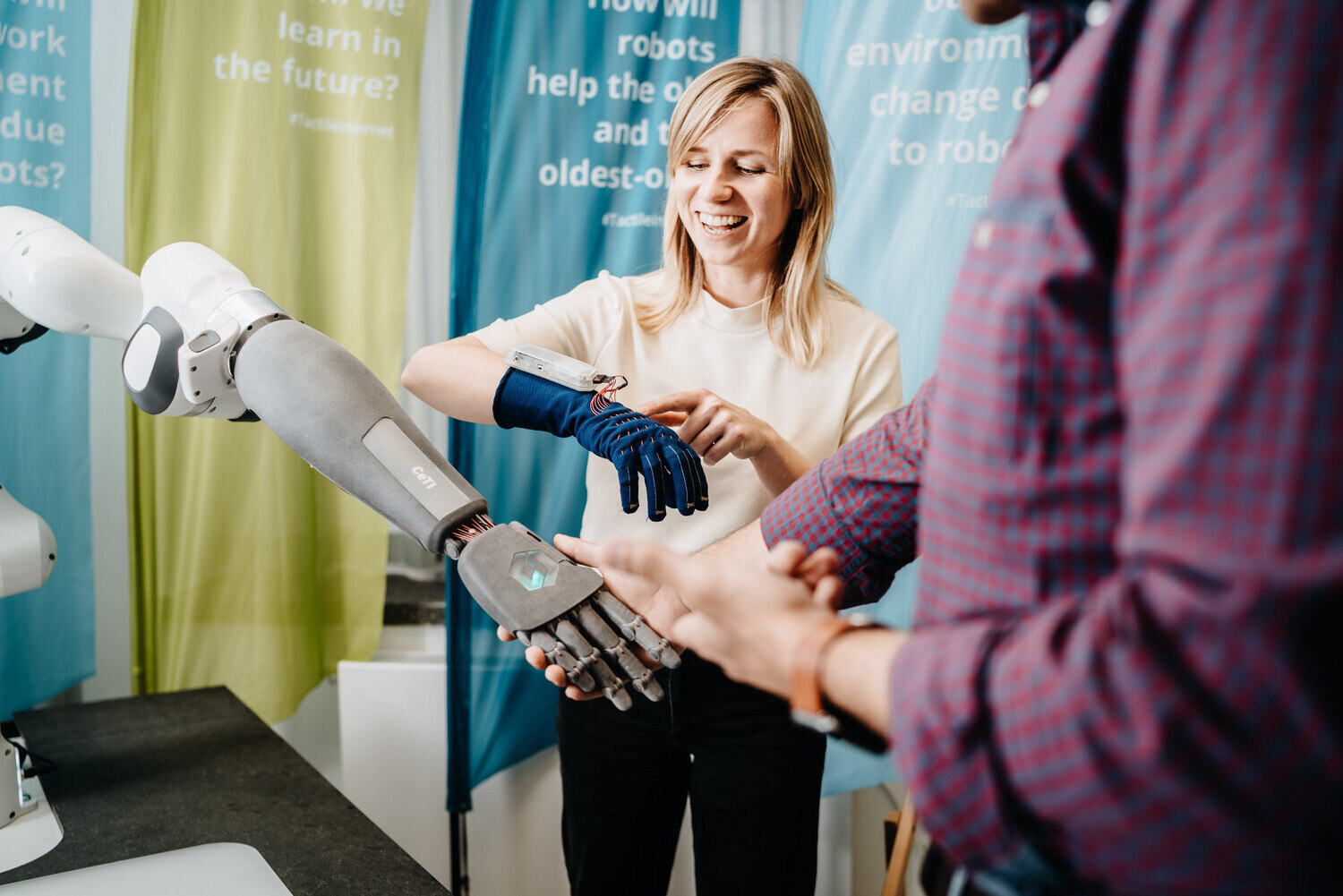 Eine Person gibt einem Roboterarm die Hand. Im Hintergrund eine Frau mit einem blauen Handschuh.